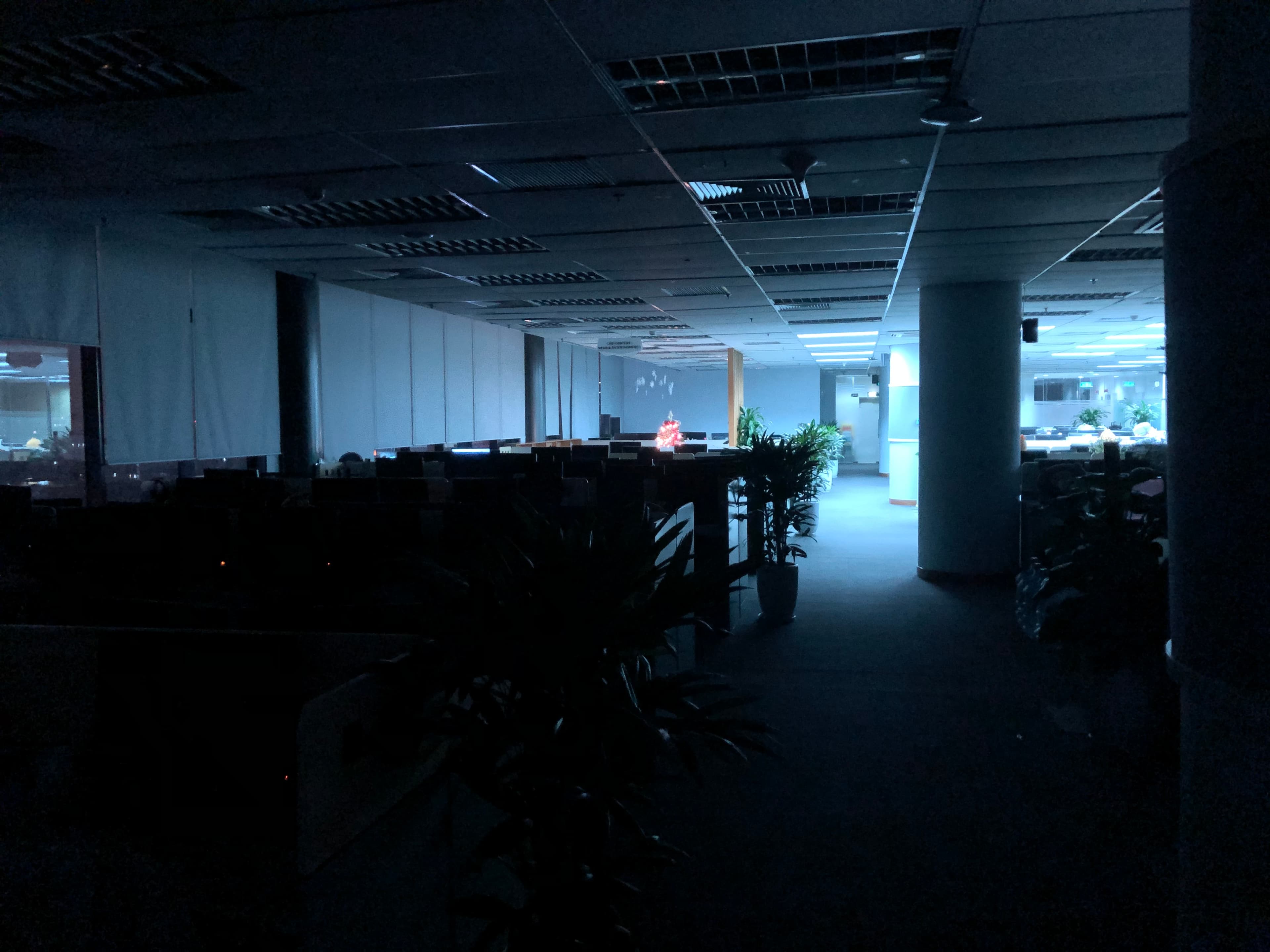Dimly lit office with empty cubicles and potted plants. A single illuminated screen is visible in the distance.